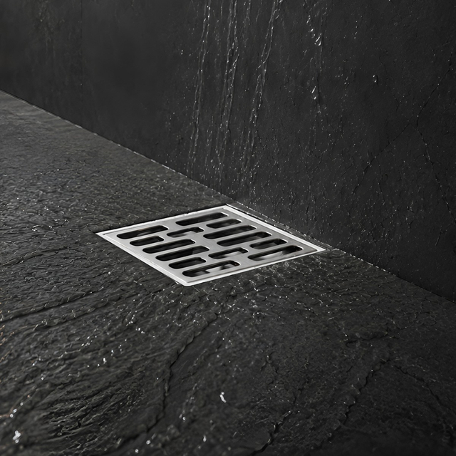 Square stainless steel bathroom floor drain set in a dark tiled shower floor with water flowing toward the grate